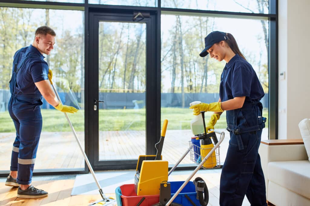 Agents de nettoyage professionnels souriants en uniforme bleu. L'homme lave le sol en bois, la femme prépare un spray près de son chariot à outils.