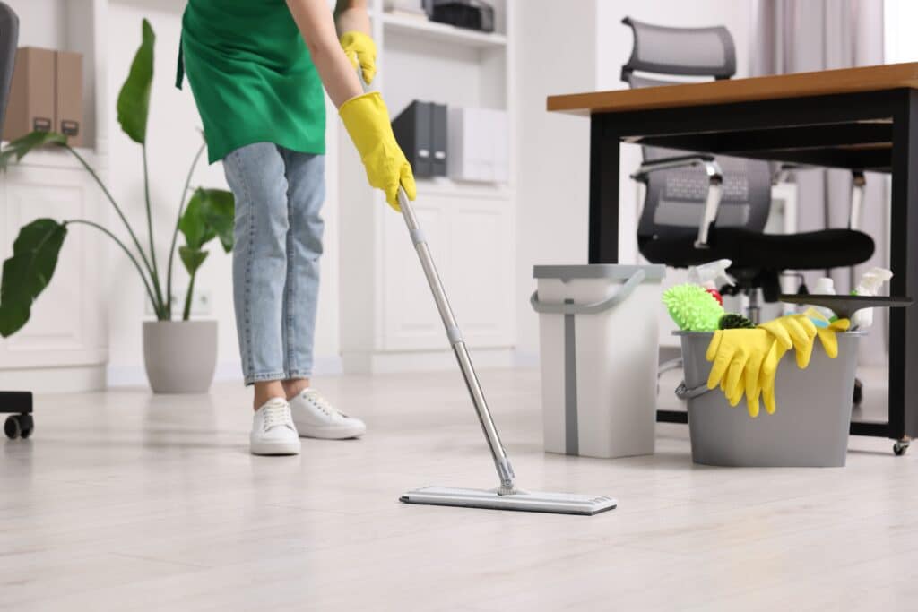 Nettoyage de bureau efficace : sol impeccable et hygiène parfaite Personne en tablier vert et gants jaunes nettoyant un sol d'office clair avec une serpillière. Seau de produits de nettoyage à côté.