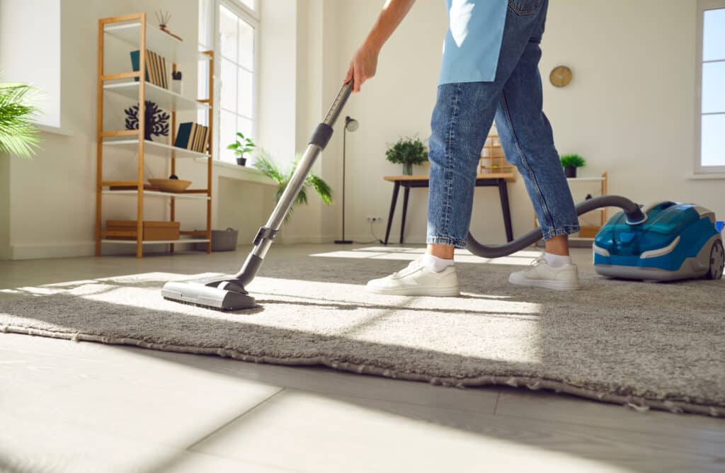 Nettoyage efficace: aspirer un tapis avec un aspirateur Personne en jean et tablier bleu aspirant un tapis beige avec un aspirateur dans un salon lumineux. Étagère en bois et fenêtre en arrière-plan.